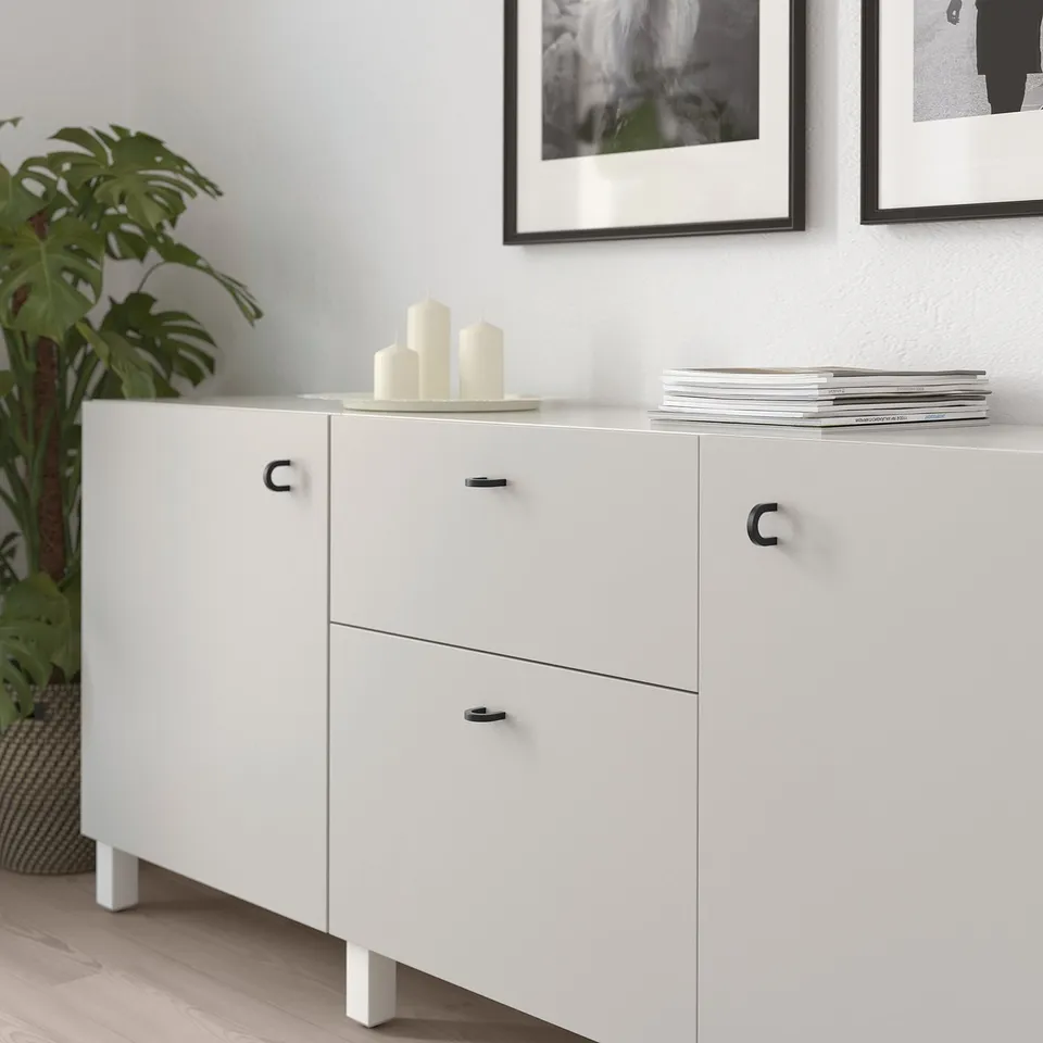White cabinet with two drawers and two doors, featuring black handles. The top holds candles and magazines.