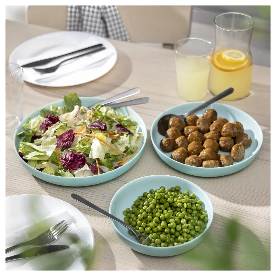Three light blue VITBLECKA bowls with salad, meatballs, peas, and cutlery on a table, showcasing a meal setup in soft turquoise tempered glass.