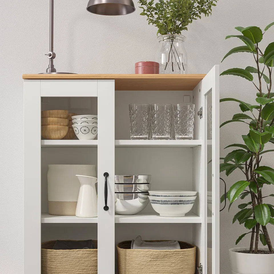 White cabinet with glass doors, displaying bowls, jars, and cups on wooden shelves, beside a tall green plant.