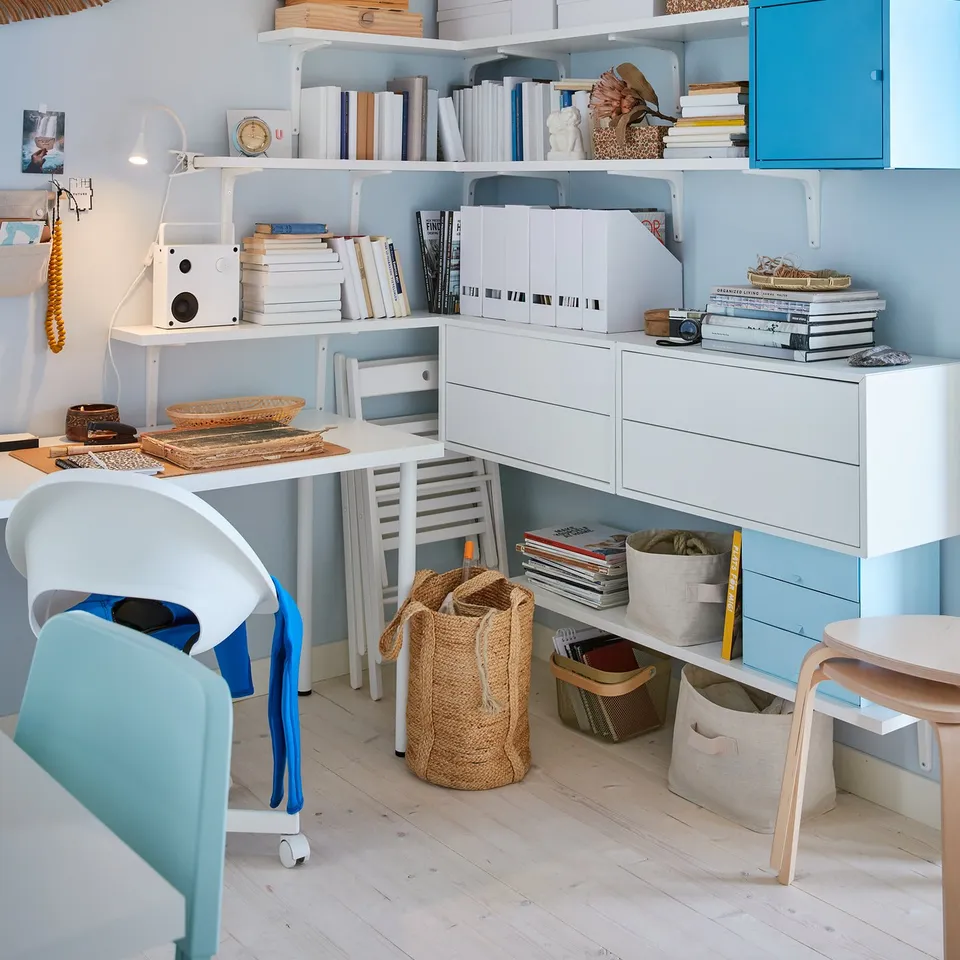 Home office with white BERGSHULT shelves holding books and storage boxes, blue cabinet, wicker baskets, desk with chair, against light blue wall.