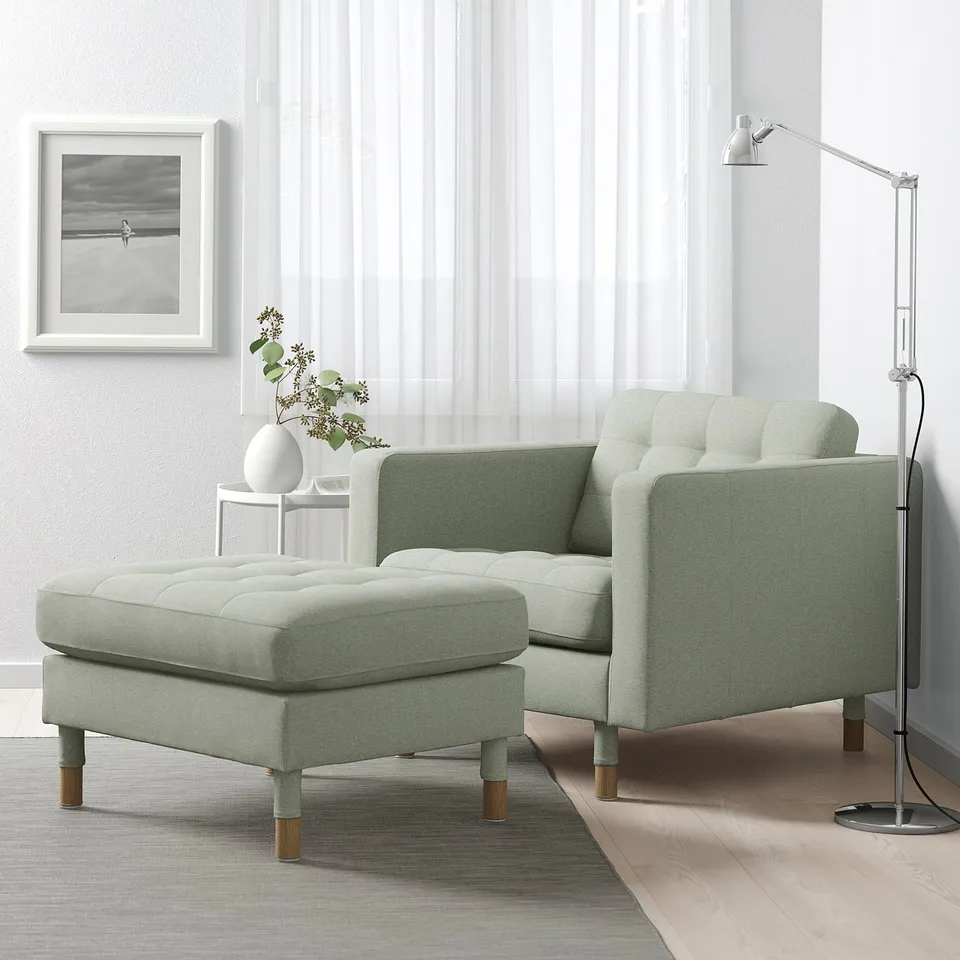 Modern living room with green sofa, footstool, lamp, vase, grey curtains, art, wood floor.