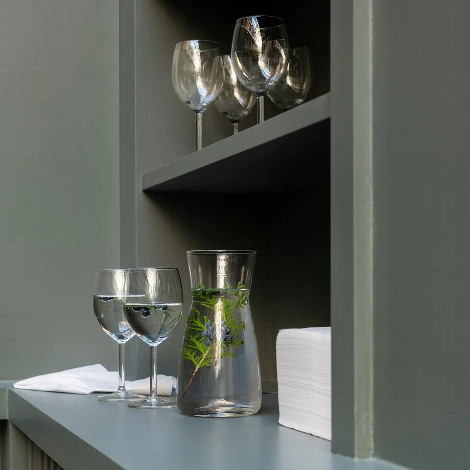 Clear glass carafe with greenery sits on grey shelf, flanked by two wine glasses, all against green backdrop.