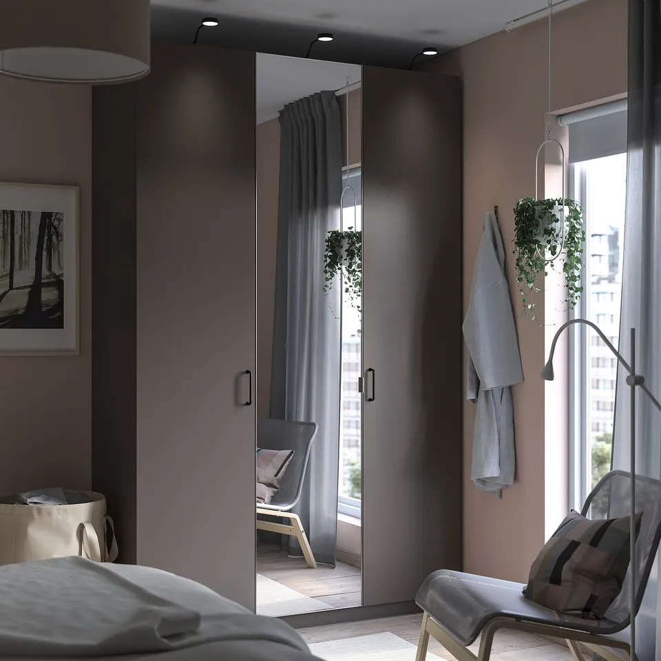 Modern bedroom with grey PAX wardrobe, mirror doors, white chair, hanging plants, large window, and soft lighting.