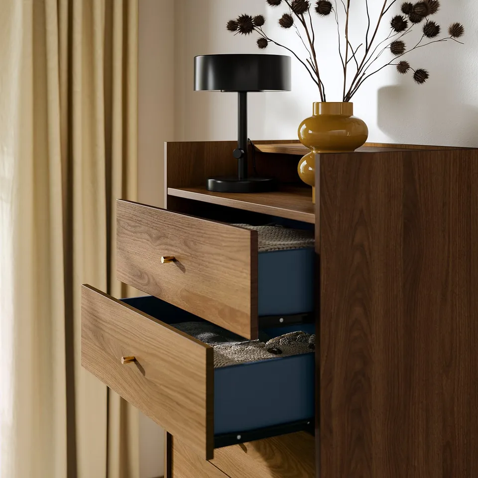 Wooden dresser with three open drawers, metal handles, lamp, and yellow vase with plant on top.