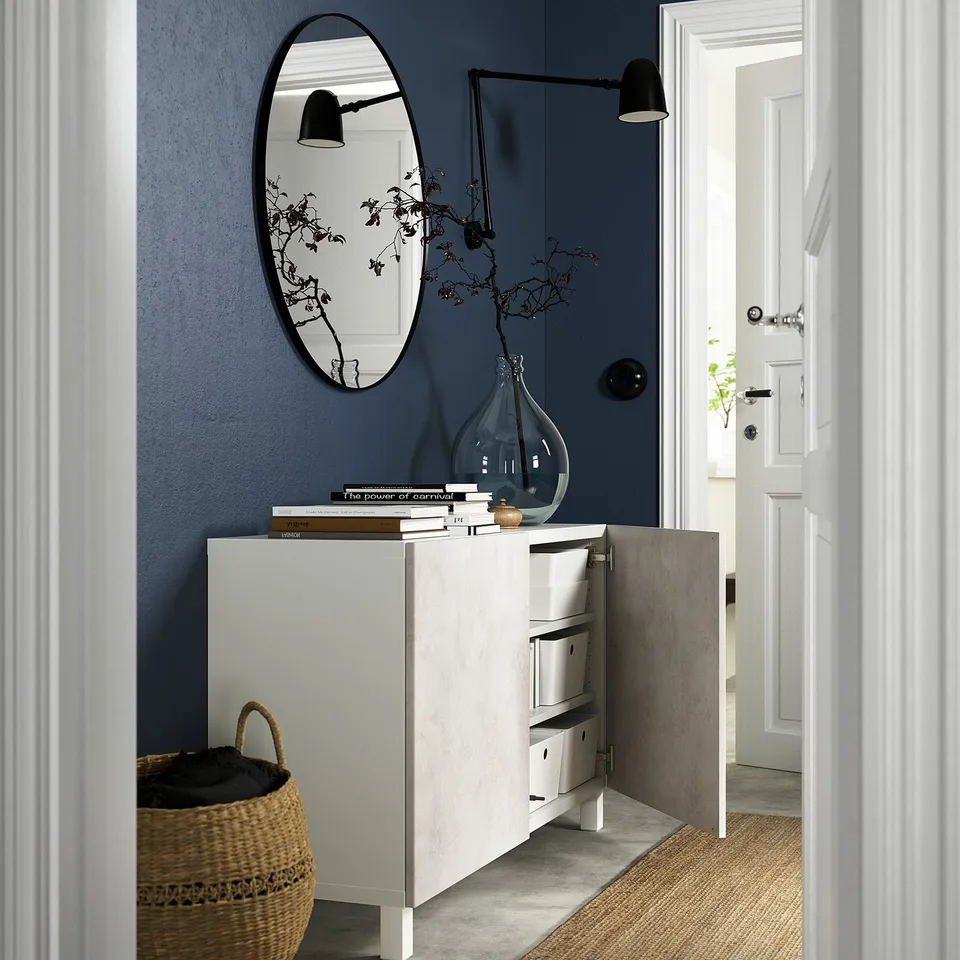 Modern entryway with white drawer unit, dark blue wall, mirror, vase with branches, and basket.
