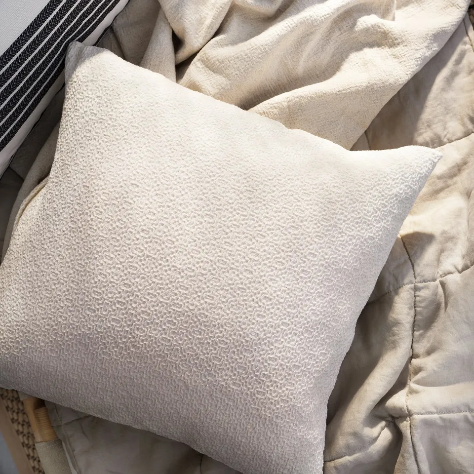 Neutral beige cushion with textured cotton cover on pile of linens.