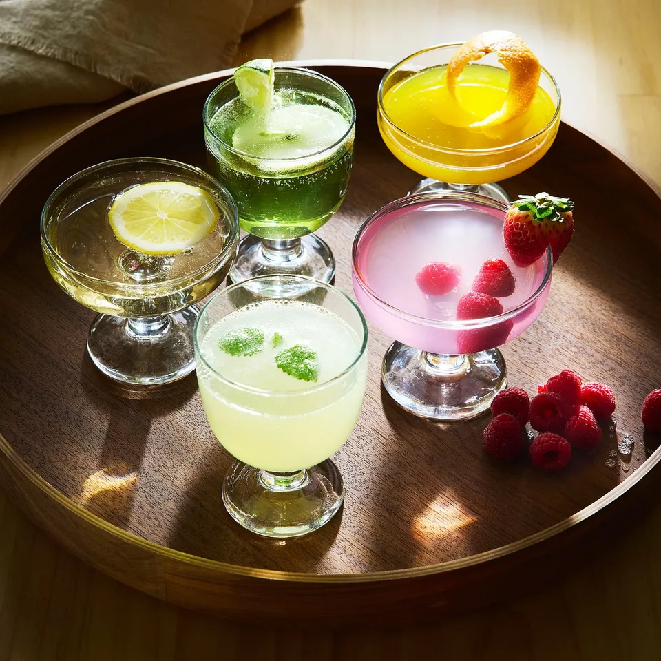 Five colourful cocktails in goblet-style glasses on a wooden tray, garnished with fruit slices and berries.