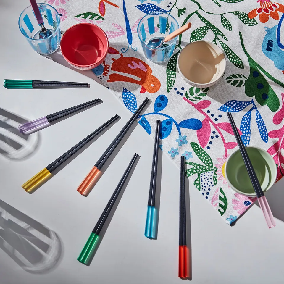 Brightly coloured chopsticks in various hues; some small ones, all laid out on playful, patterned tablecloth.