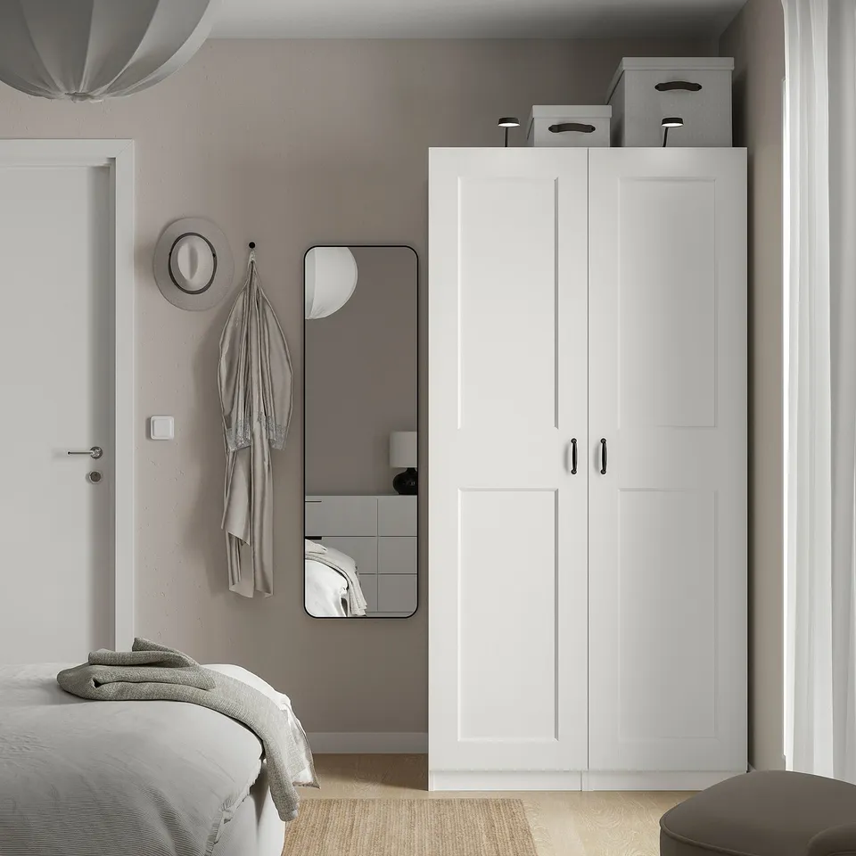 Modern bedroom with white PAX wardrobe, bed, mirror, and grey hat on hooks. Minimalist design, clean lines.