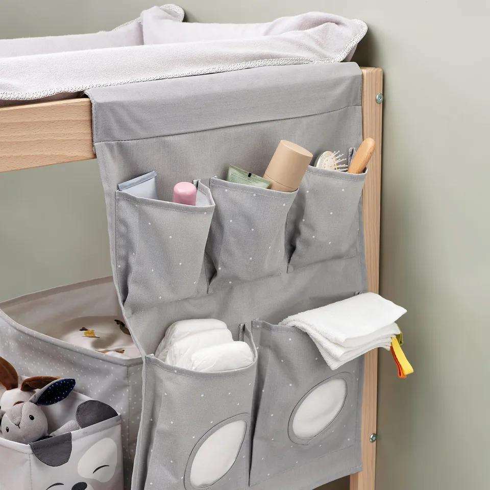 Grey hanging organiser with multiple pockets holding baby toiletries; attached to wooden furniture.