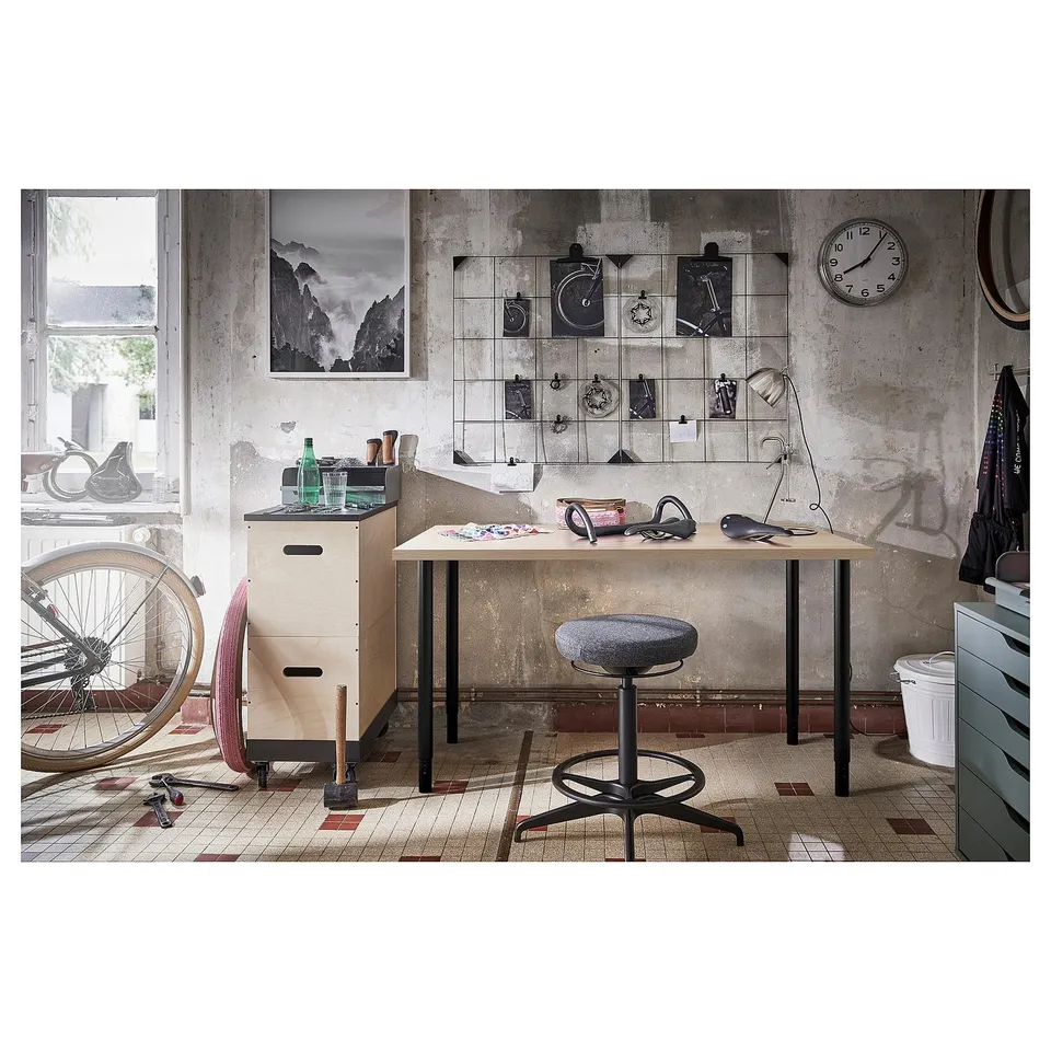 Industrial-themed workspace with LAGKAPTEN table, bike artwork, and clock on concrete wall; desk has grey chair and wooden cabinet with tools.