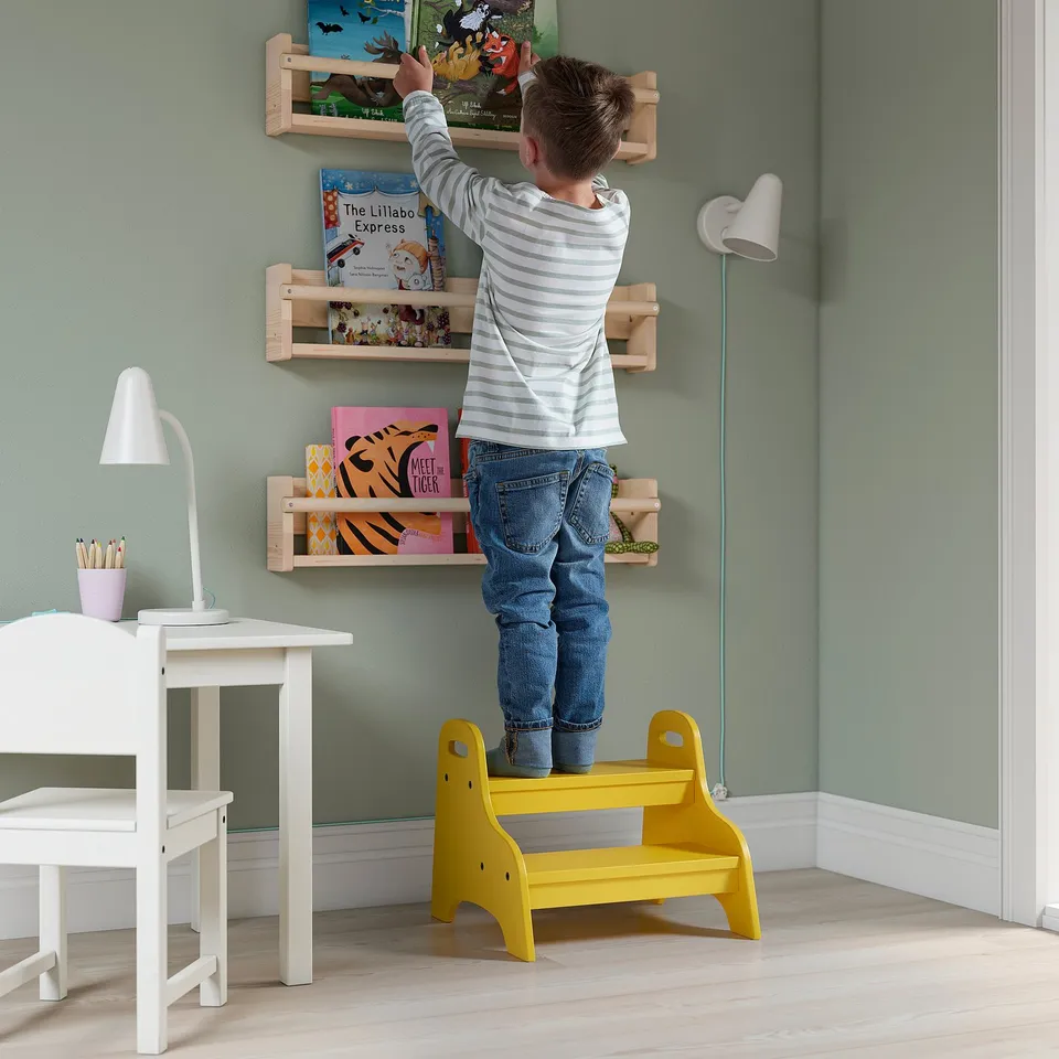 Child standing on yellow TROGEN stool, reaching for books on wall shelves. White desk and lamp nearby.