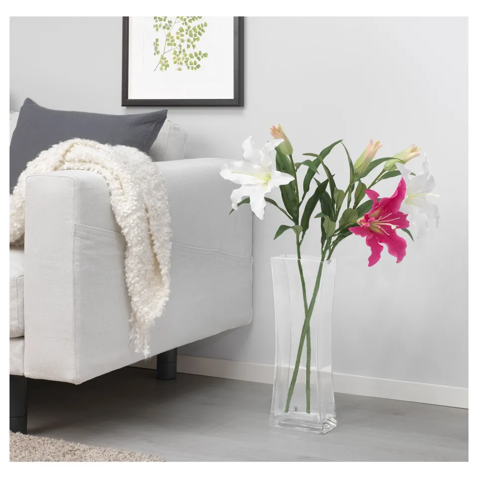 A modern living room with a white sofa and a clear glass vase holding artificial lilies, white and hot pink, near the sofa on a light wooden floor.