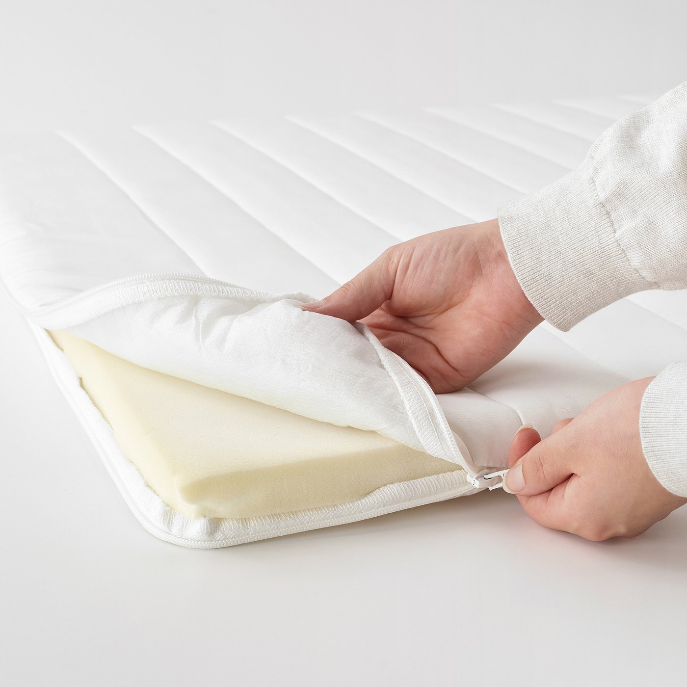 A person is attaching a white mattress pad with foam cushioning, shown unfurling and securing it using a clip.