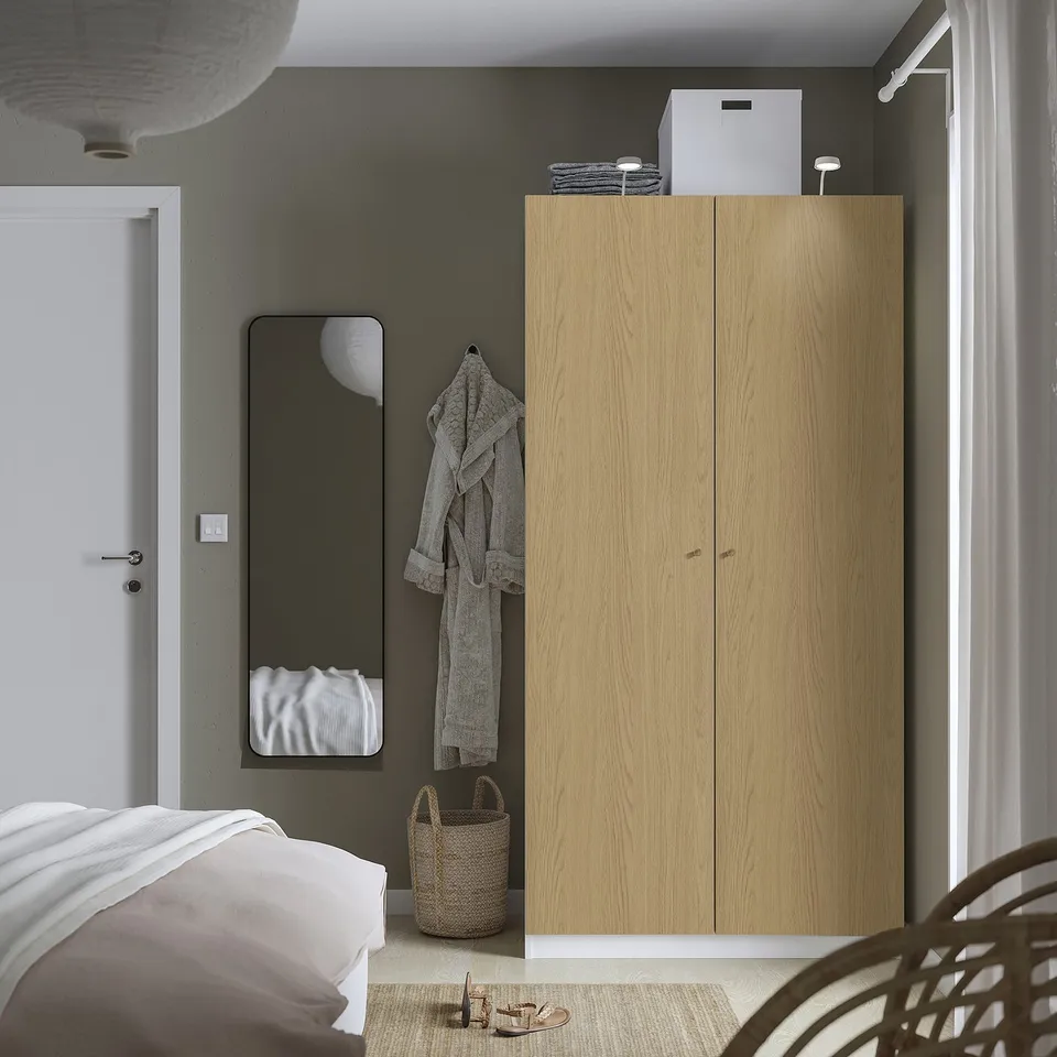 Minimalist bedroom with tall wood PAX wardrobe, wicker basket, grey robe, black mirror, white door, and bed with white linens.