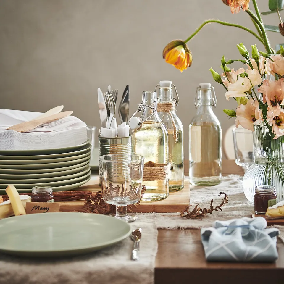 A rustic table set with green FÄRGKLAR stoneware plates, glass dispensers, cutlery, and a vase of flowers.