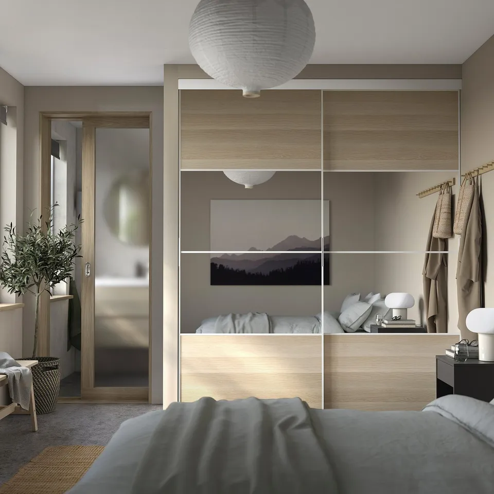 Modern bedroom with wooden built-ins, clothes rack, mountain picture, pendant lamp, grey bed, mirror, plant, and basket in minimalist decor.