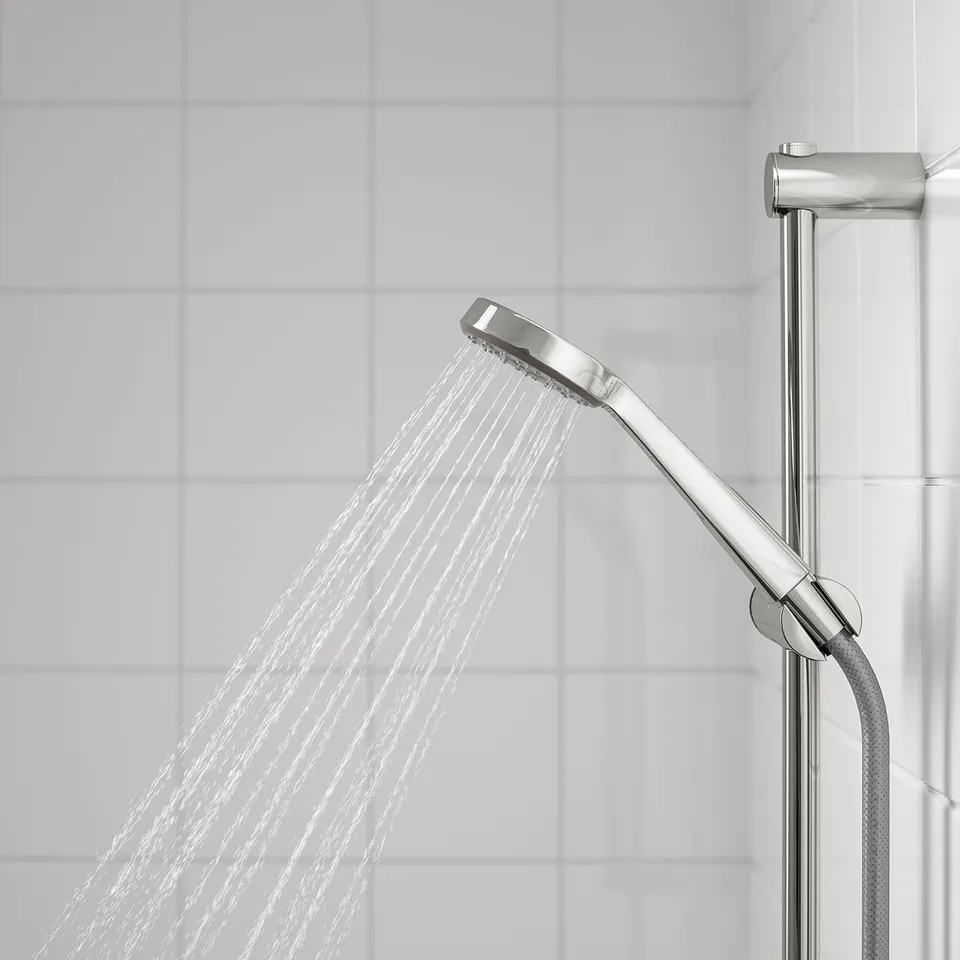 A modern chrome hand shower with full spray function operates, mounted on a holder, with water flowing in a smooth arc against white tiles.