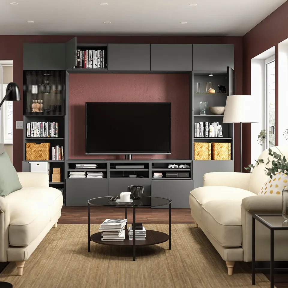 Modern living room with BESTÅ tv unit, white sofas, wooden tables, and a large flat-screen tv. Red wall, grey shelves, and stylish decor.