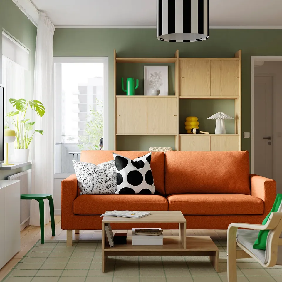 Bright living room: orange SALTSJÖBADEN sofa, colourful cushions, wooden coffee table, green plant, light shelves, striped rug, and modern lighting.