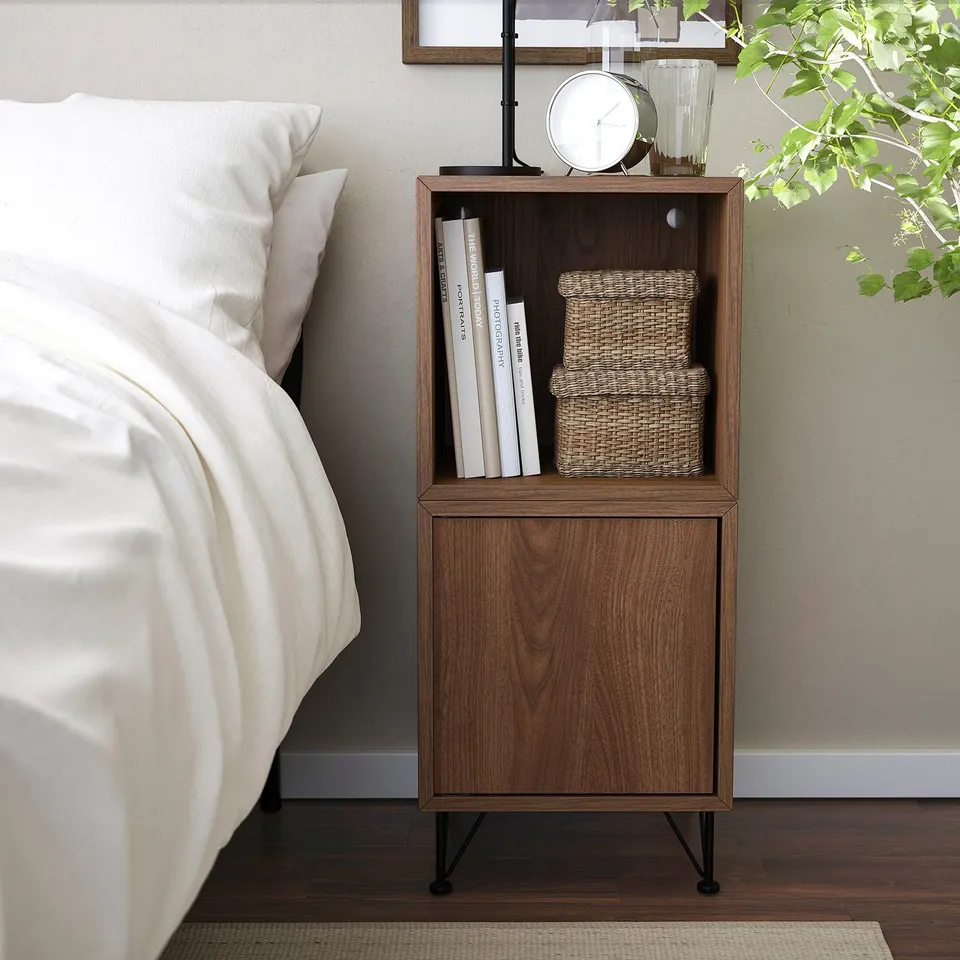 Bedside scene with wooden EKET shelf. Clock, books, and baskets displayed. White bedding beside it. Shelving tall, narrow, fits neatly in room.