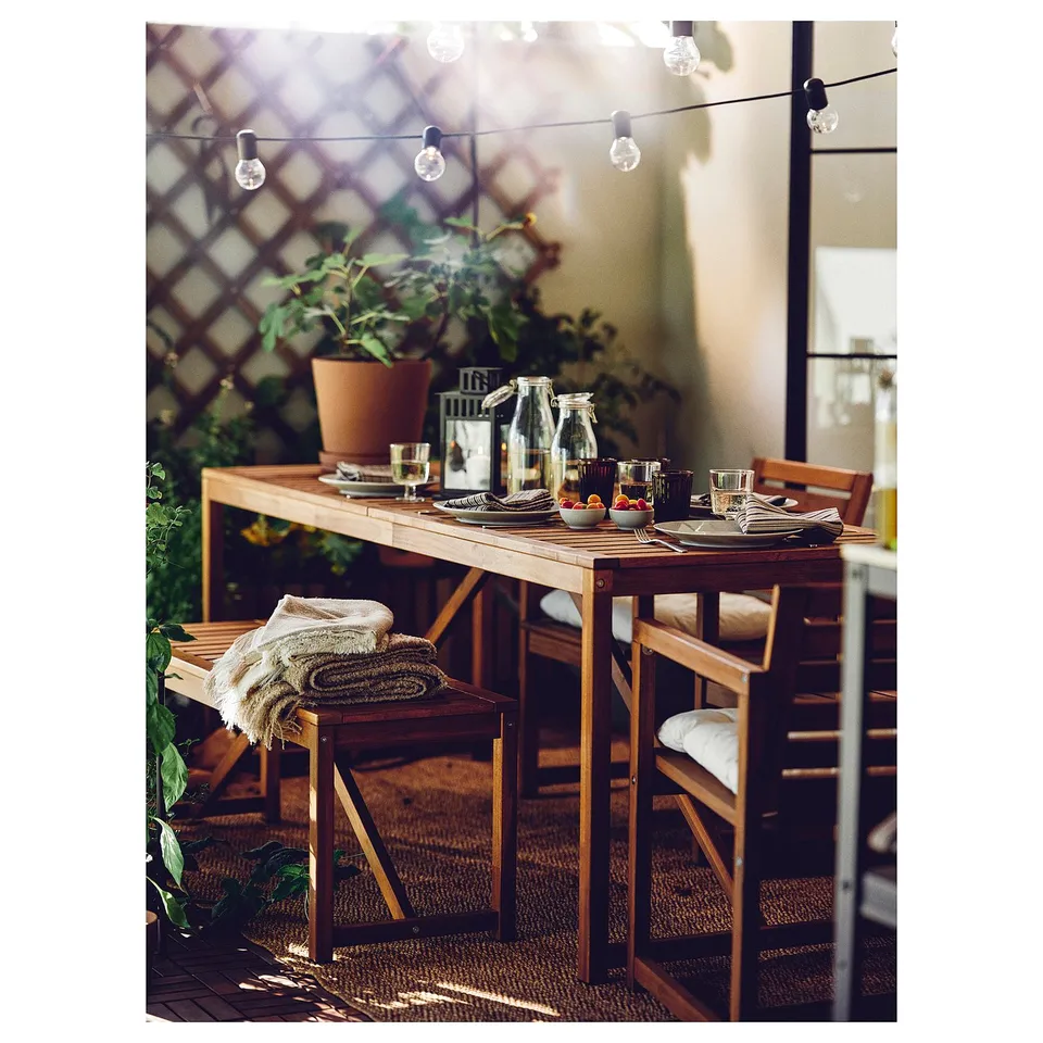 Outdoor dining setup with a brown wooden table, chairs, and string lights. Plates, glasses, and a potted plant.
