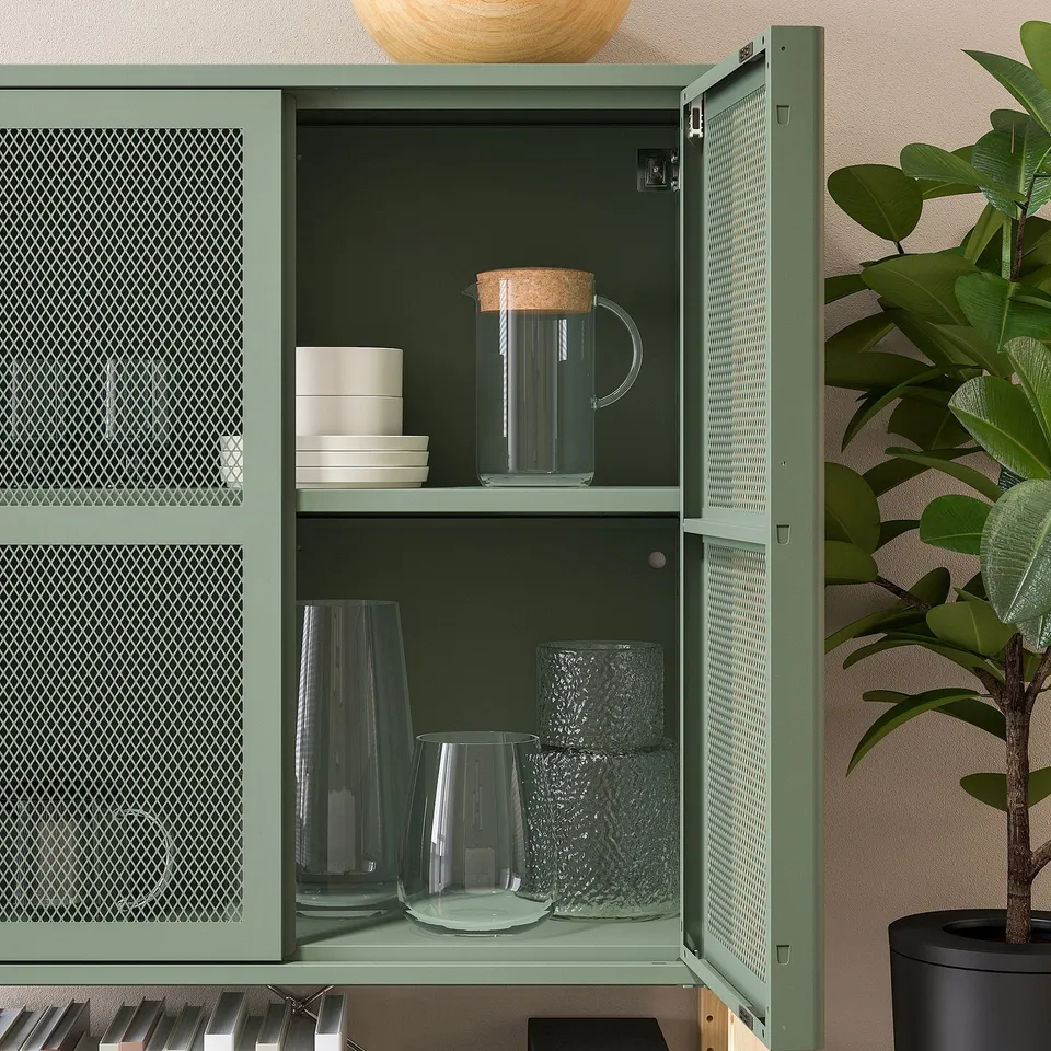 Green IVAR cabinet with glass door displaying stacked glassware and pitchers, secured to wall.