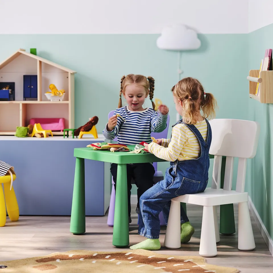 Two girls playing at green MAMMUT kids table with white chairs, turquoise wall, dollhouse, toys.