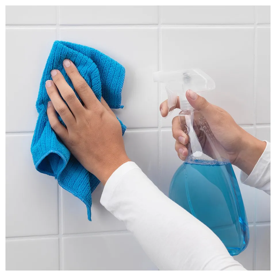 Person holding blue spray bottle and cloth, preparing to clean tiled wall with TISKEN suction holder.