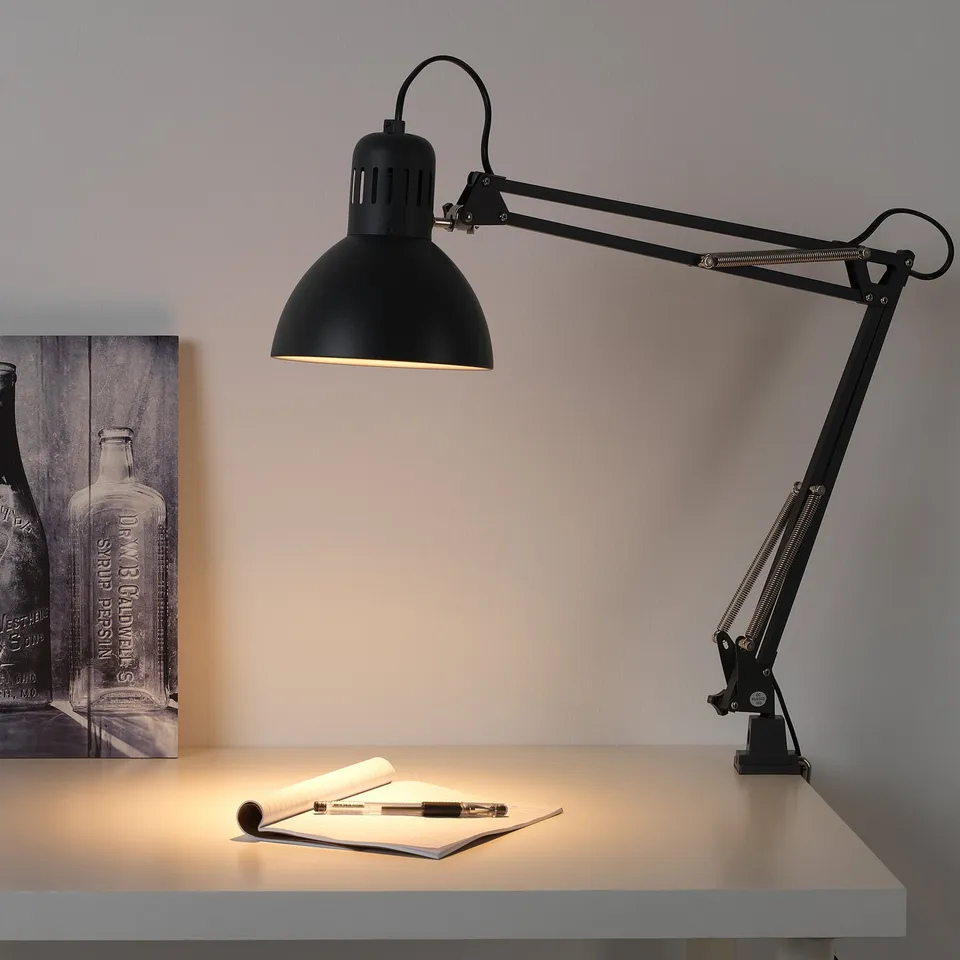 Black adjustable desk lamp illuminates a notebook and pen; classic design with flexible arm and head.