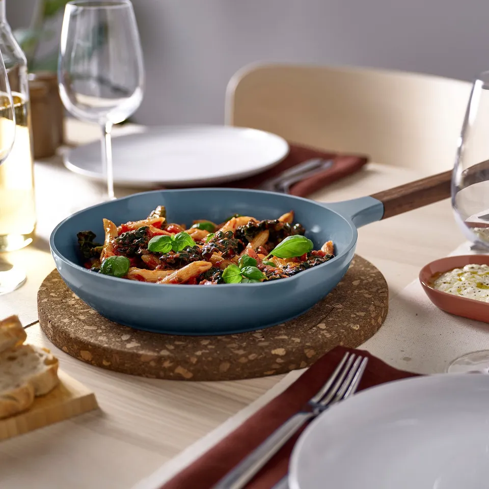 Blue cast aluminium pan with wood handles on trivet, filled with veggies, plates, glasses, bread.