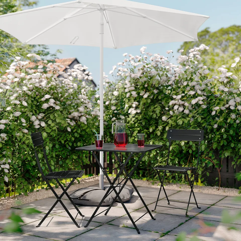 Outdoor setting with black metal table and chairs, white umbrella, red drinks. Beautiful garden view.