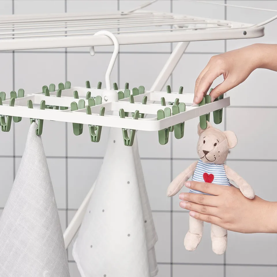 A hand hangs clothes using a white plastic drying rack with 24 green clips. A bear stuffed toy is held nearby.