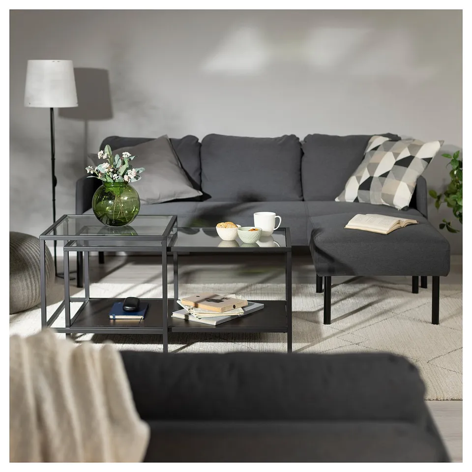 Modern living room with grey GLOSTAD sofa, glass coffee table, and grey footstool. Decorative pillow, lamp, and vase with flowers.