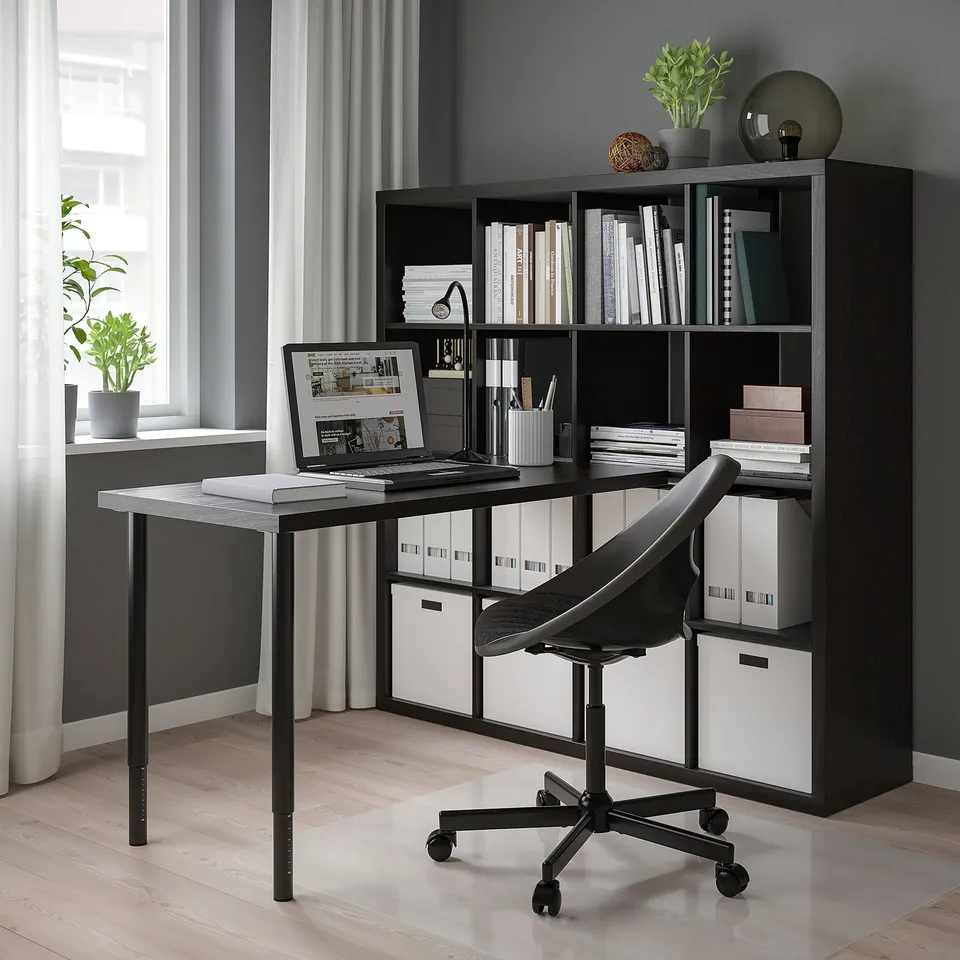 Modern home office setup with KALLAX black shelving unit, black desk, and chair. Shelves hold books, files, and plants. Laptop on desk by window.