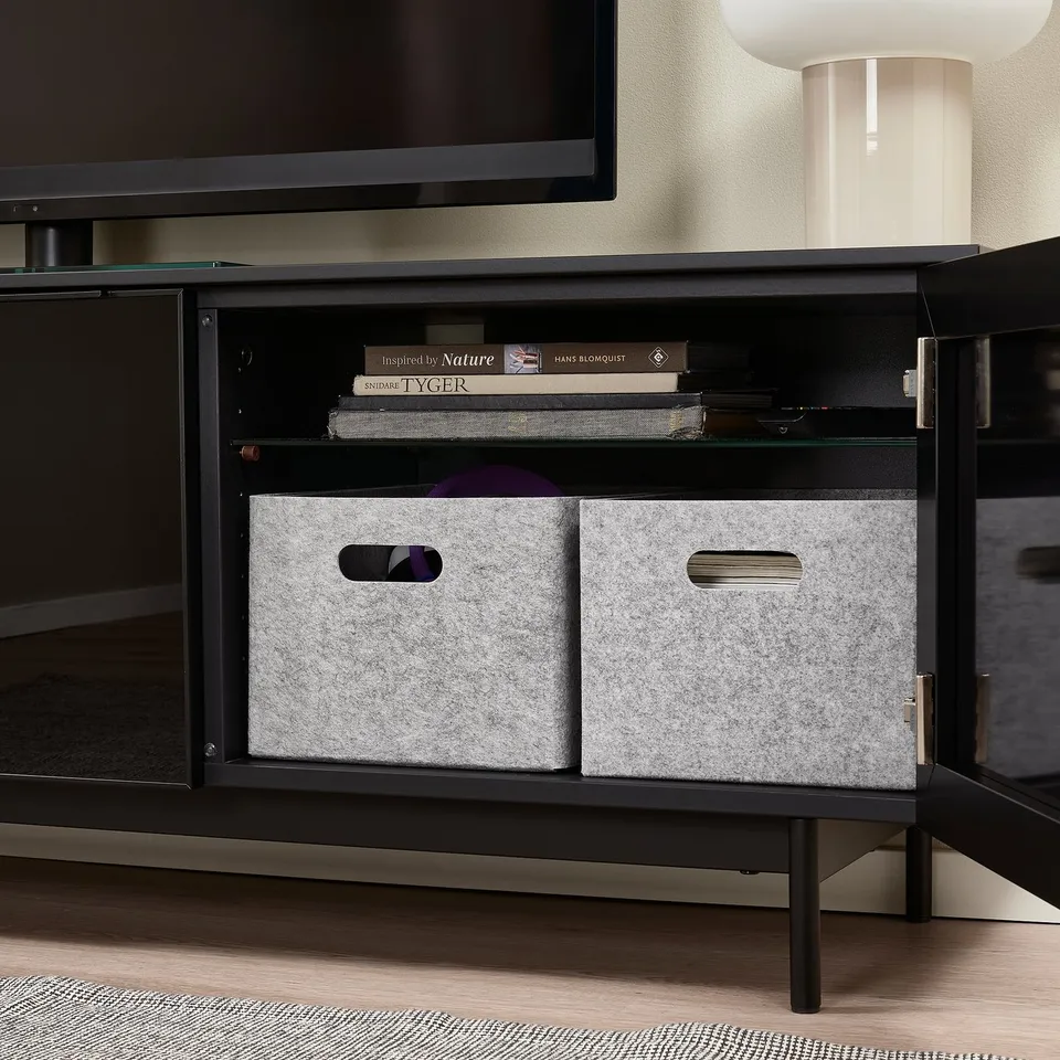 Black tv stand with glass shelf holding books and two grey felt storage boxes with cut-out handles for organisation.