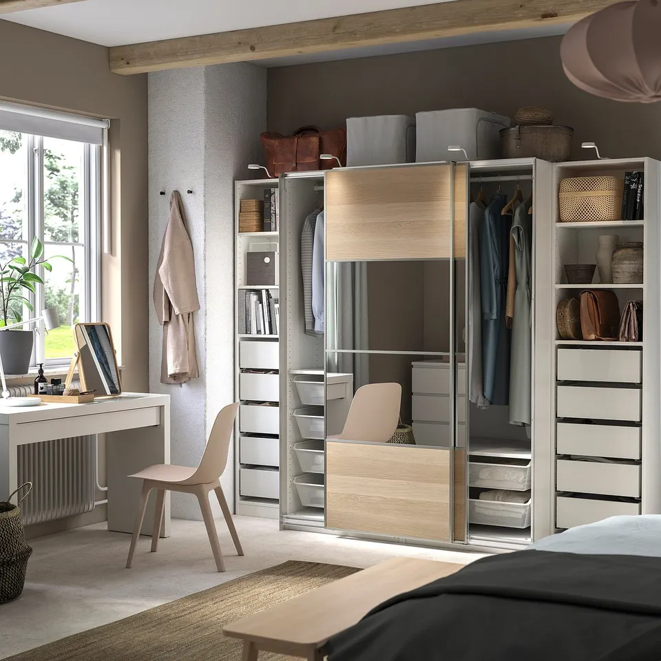 Modern bedroom with light wood PAX wardrobe, featuring mirrored slides, shelves, and drawers. Desk, chair, and rug complete the organised space.