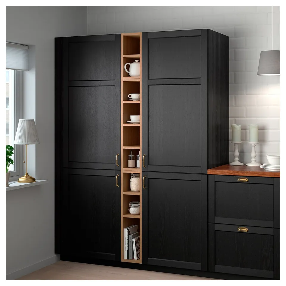 Black VADHOLMA kitchen storage unit with dark wood, open section, teacups, jars, books, near window, gold lamp.