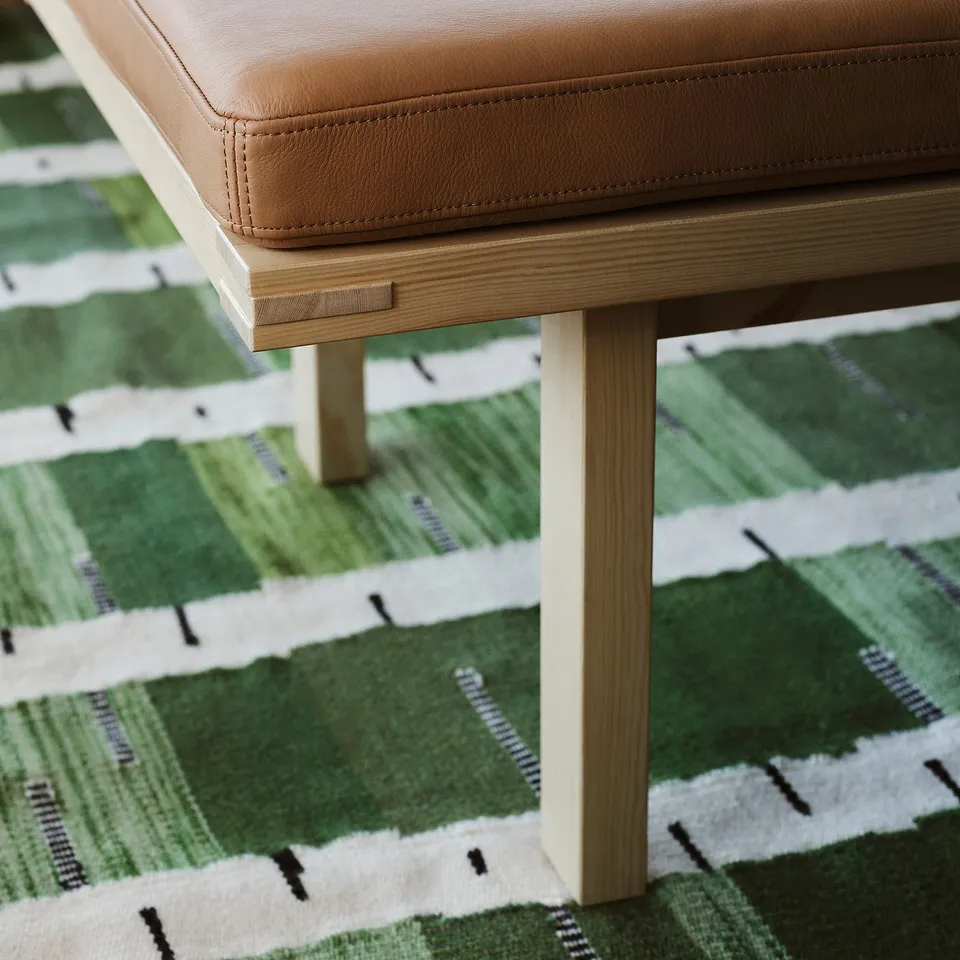 A handwoven green rug with a birch tree pattern, placed under a brown leather stool with wooden legs.