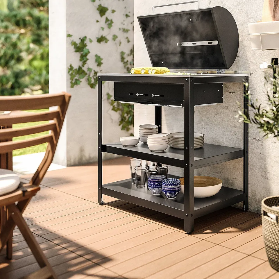 Outdoor bbq setup with GRILLSKÄR charcoal grill, black, on tiled patio. Grill contains food; shelves hold plates and cups.