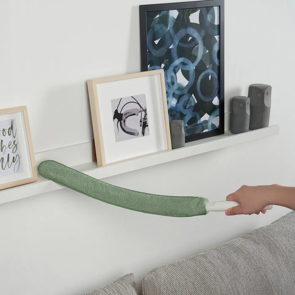 A person uses a PEPPRIG microfiber duster to clean a low wall shelf. The duster has a replaceable cover and is held securely.