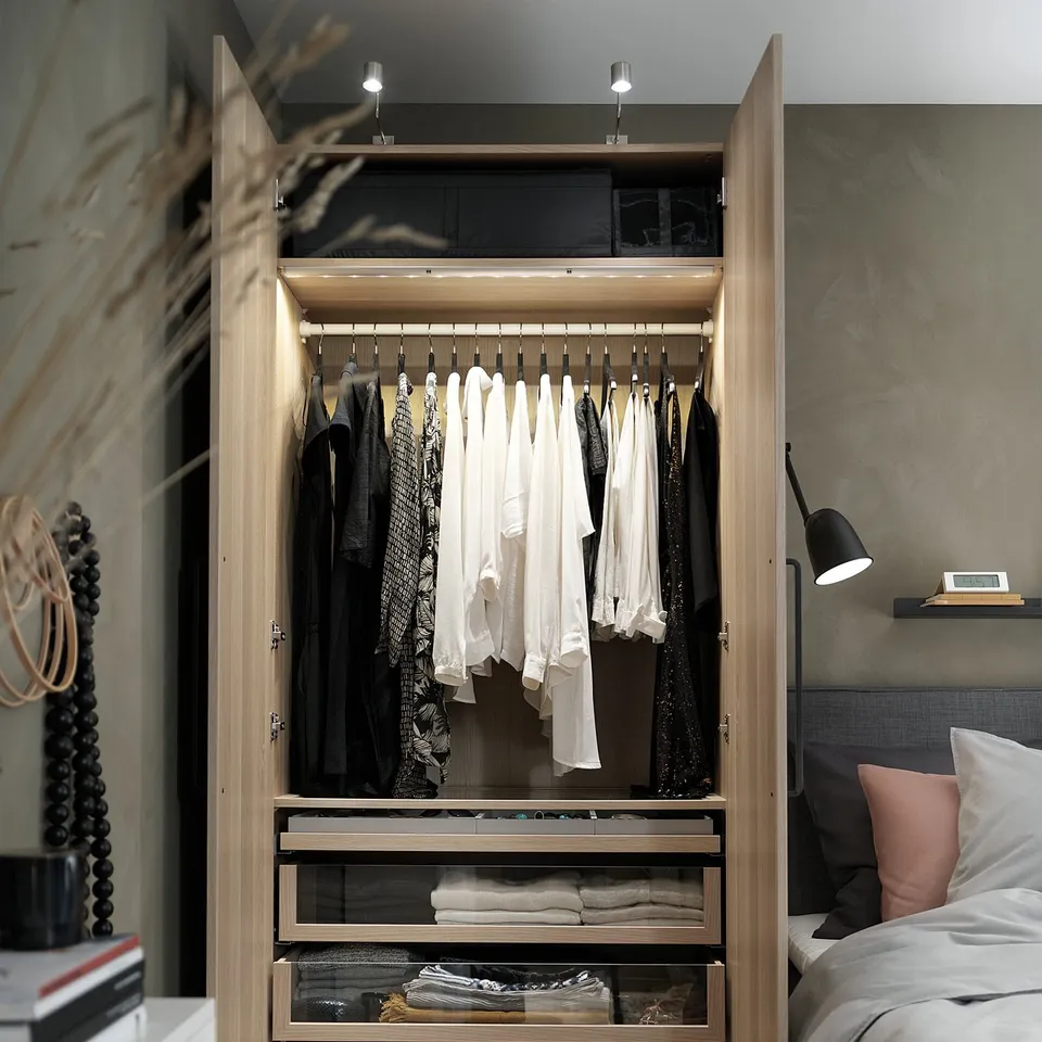 An open wooden closet, PAX, displays clothes and folded items; it has glass drawers, shelves, and led lighting.