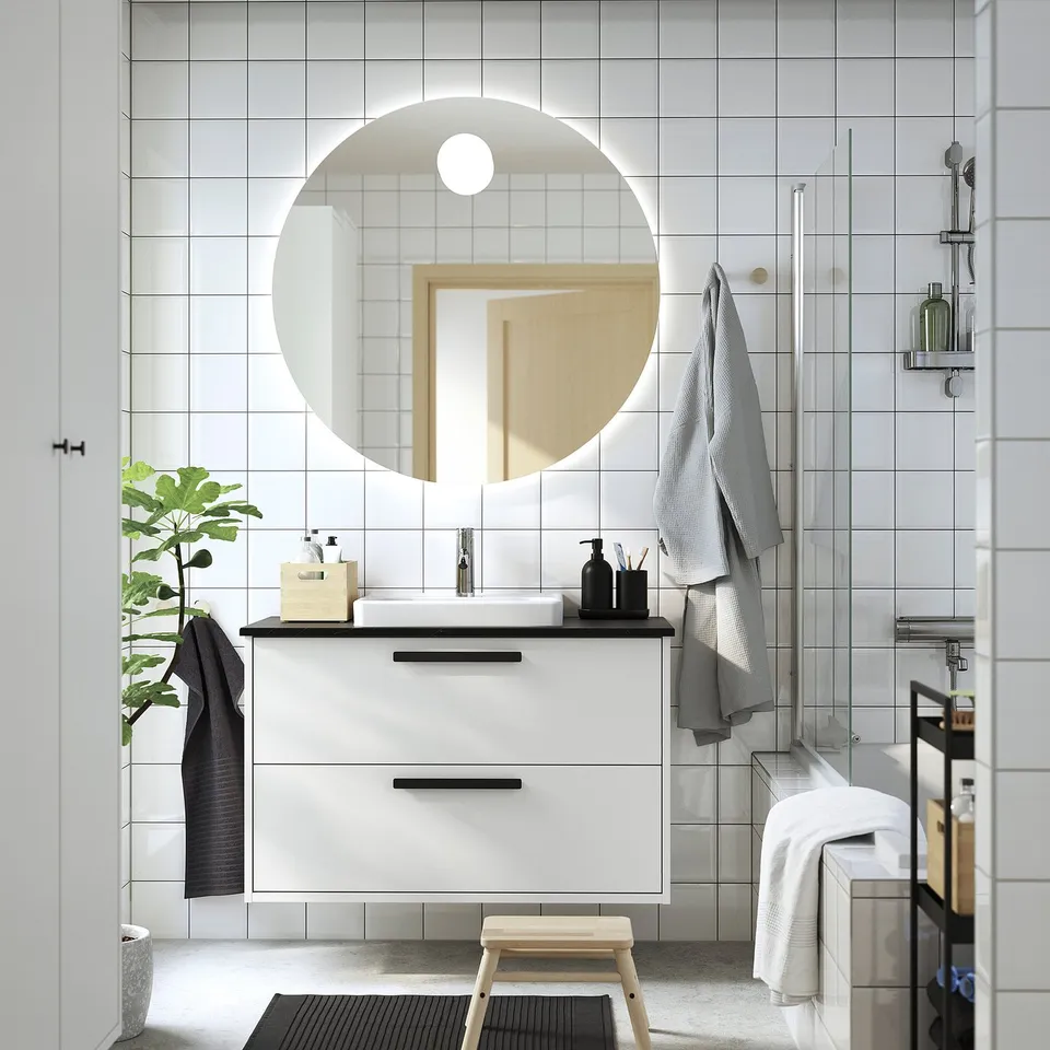 Modern bathroom with TOLKEN countertop, mirror, towels, stool, plant. Black marble effect contrasts white cabinet.