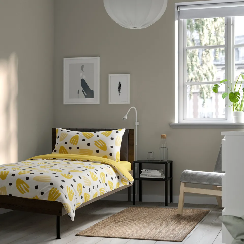 Bright bedroom with yellow-patterned bedding, grey walls, large window, and wall art. The bed is the centrepiece, topped with a colourful bedspread.