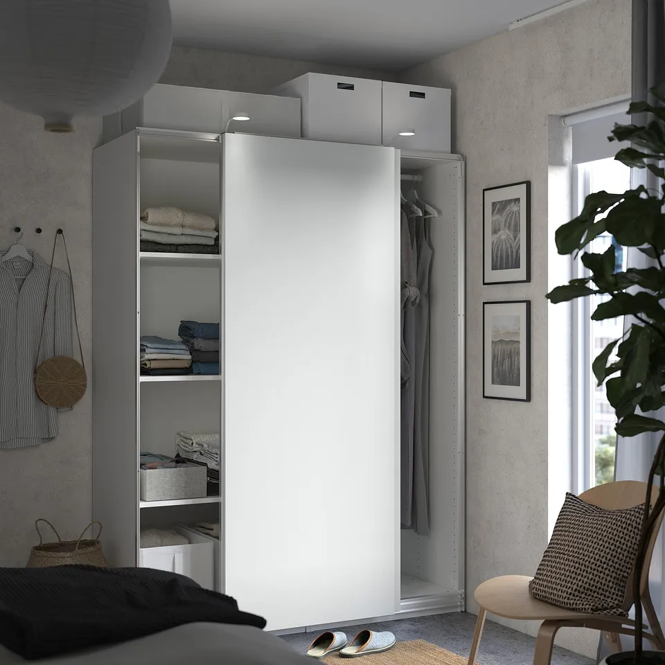 A modern grey bedroom featuring a white sliding door PAX wardrobe with shelves and hanging space, complemented by wall art and a wooden chair.