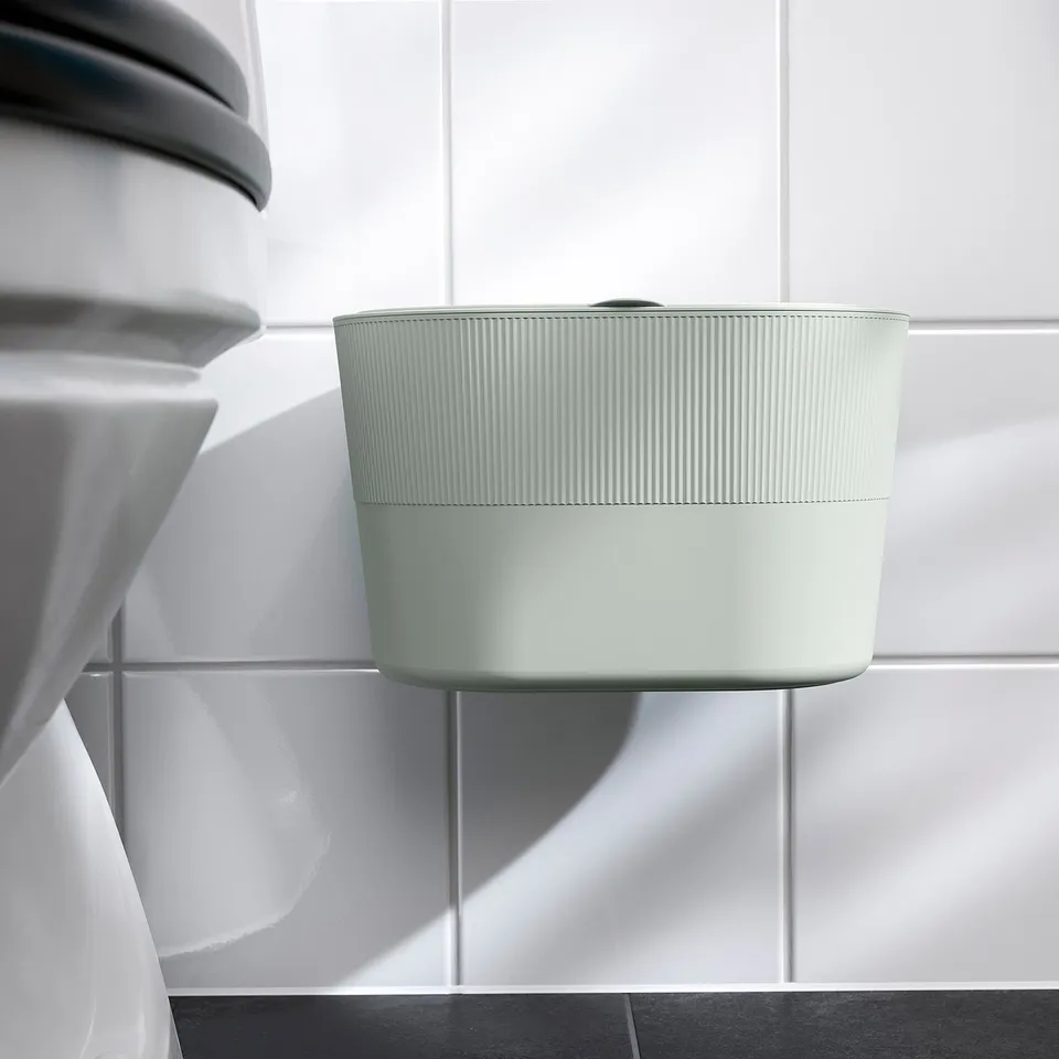 Light green waste bin with lid hangs on tiled wall, next to a white appliance.