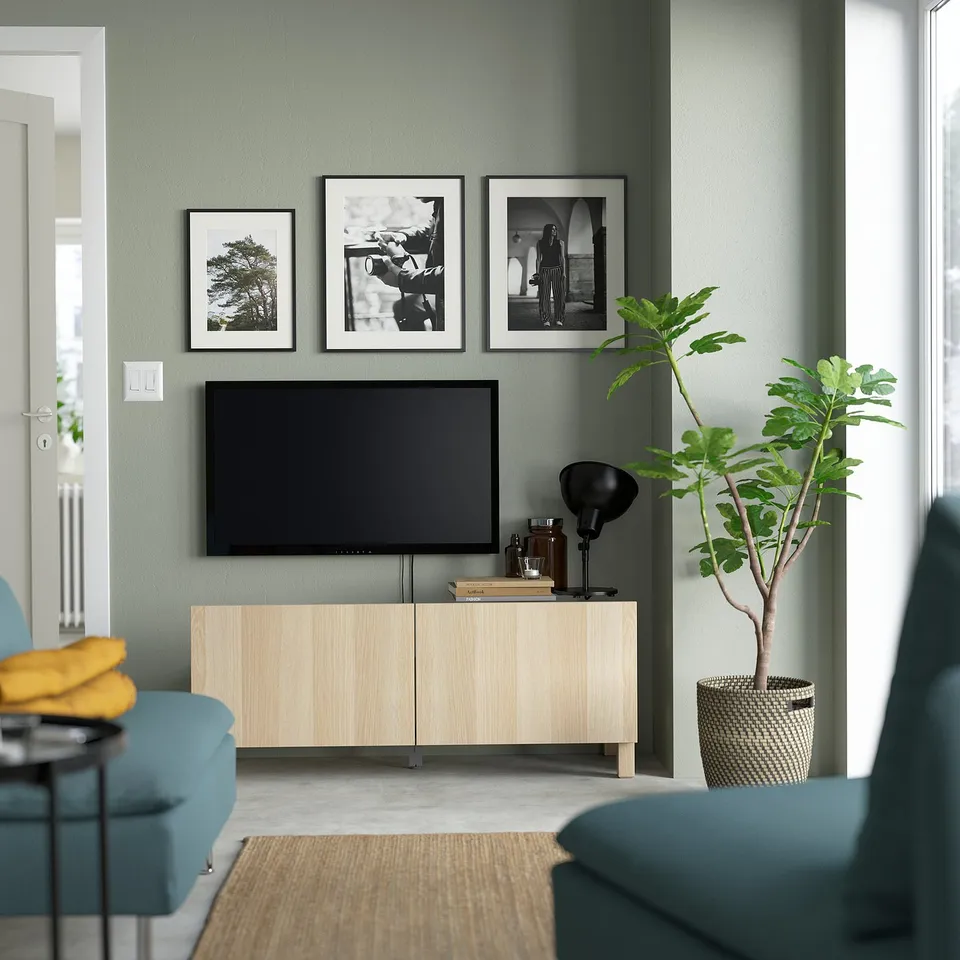 Modern living room with flat-screen tv, books, lamp, black-and-white photos, green wall, plant, teal sofa, yellow pillow.