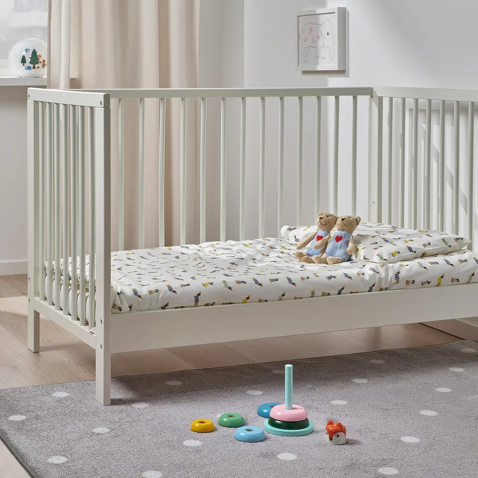 Small white wooden baby crib with a patterned mattress and two stuffed animals, surrounded by colourful stacking rings toy on a polka dot rug.