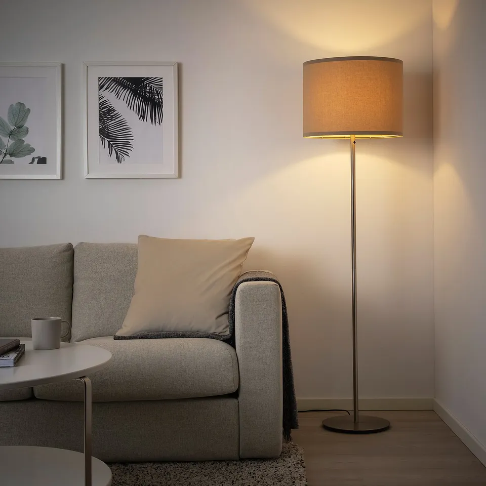 A cosy living room with a grey, modern SKAFTET floor lamp beside a light grey couch. The lamp has a beige lampshade, adding warmth to the space.