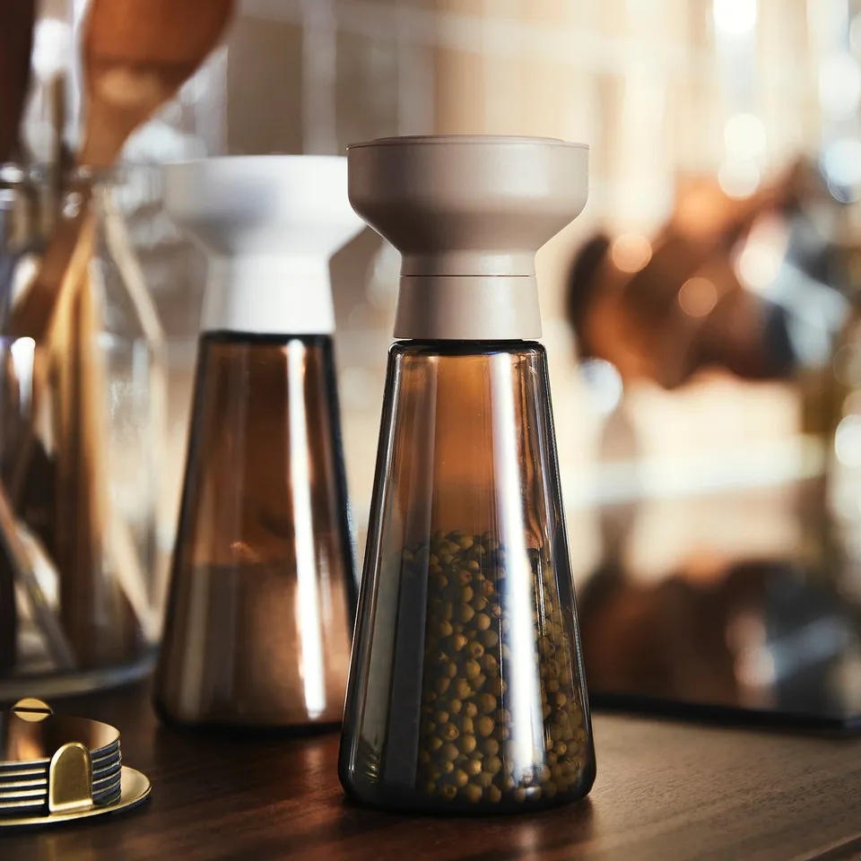 Two modern spice mills on a wooden table with a blurry restaurant background. One contains green peppercorns.